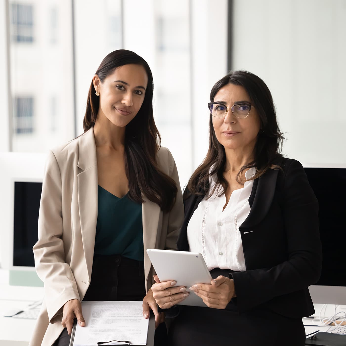 A young professional woman and senior businesswoman in glasses posing in office for portrait, standing close together, looking at camera. A young professional woman and senior businesswoman in glasses posing in office for portrait, standing close together, looking at camera.