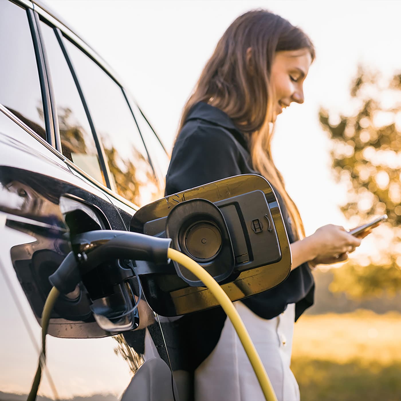 A woman charging her EV vehicle while looking at her phone. A woman charging her EV vehicle while looking at her phone.